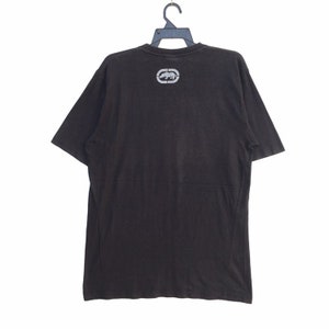 May include: Black short-sleeved t-shirt with a crew neck. The shirt features a silver logo of a rhinoceros inside an oval on the upper back. The shirt is hanging on a black hanger against a white background.