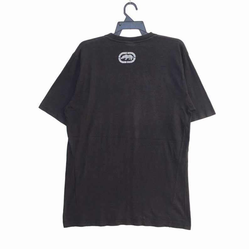 May include: Black short-sleeved t-shirt with a crew neck. The shirt features a silver logo of a rhinoceros inside an oval on the upper back. The shirt is hanging on a black hanger against a white background.