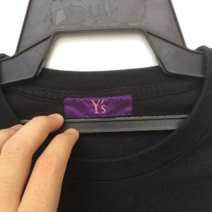 May include: Black crew neck t-shirt with a purple label that reads "Y's" in pink lettering. The shirt is hanging on a black hanger. The image is a close-up of the shirt's neckline.