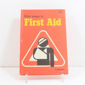 May include: A red book with the title "First steps in First Aid" and a black and white illustration of a person with a broken arm inside a red triangle. The book has the word "Ladybird" printed on the bottom.