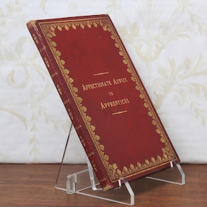 May include: A vintage book titled "Affectionate Advice to Apprentices" is displayed on a clear acrylic stand. The book has a deep red cover with gold embossed decorative borders and lettering. The book is slightly angled, showcasing its title.