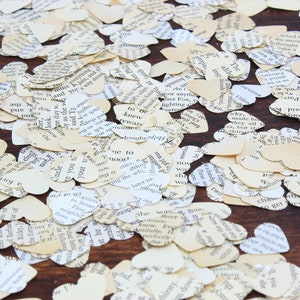May include: A close-up image of a large quantity of heart-shaped confetti made from recycled book pages. The confetti is scattered on a brown wooden surface.