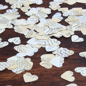 May include: A close-up of a wooden surface covered in white and cream paper heart confetti. The hearts are cut from pages of old books and have black text printed on them.