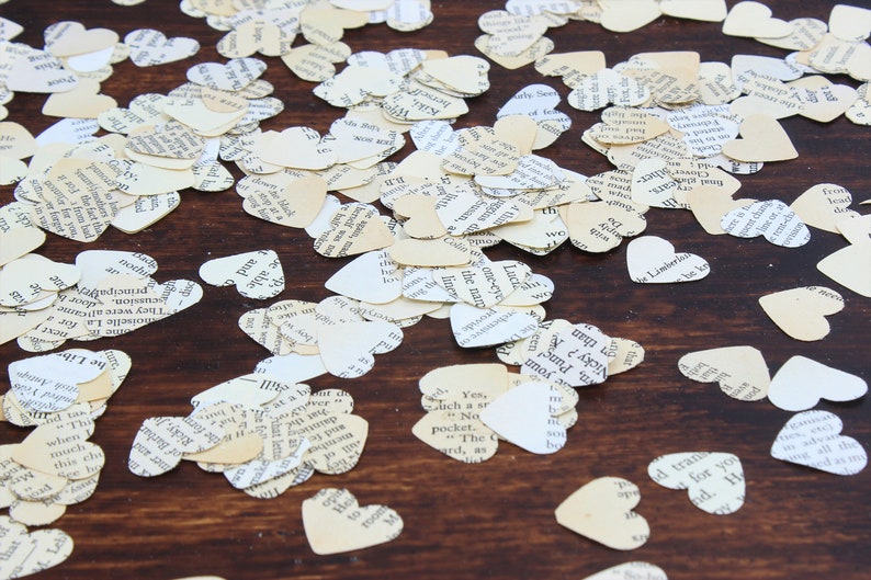 May include: A close-up of a wooden surface covered in white and cream paper heart confetti. The hearts are cut from pages of old books and have black text printed on them.