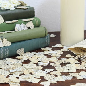 May include: A stack of vintage green hardcover books with a pile of paper heart confetti scattered around them. The confetti is made of old book pages and is a light cream color. A white vase is to the right of the books.