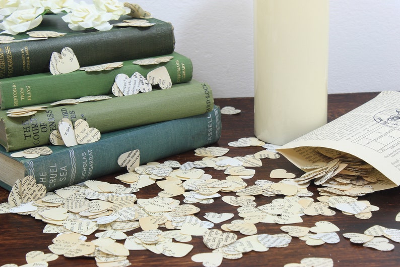 May include: A stack of vintage green hardcover books with a pile of paper heart confetti scattered around them. The confetti is made of old book pages and is a light cream color. A white vase is to the right of the books.