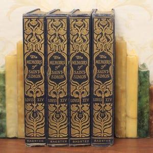 Puede incluir: Cuatro libros antiguos titulados "The Memoirs of Saint-Simon" con letras doradas y diseños ornamentados en cubiertas azul marino. Las lomos muestran los números de volumen I, II, III y IV. Los libros están flanqueados por sujetalibros amarillos y verdes.