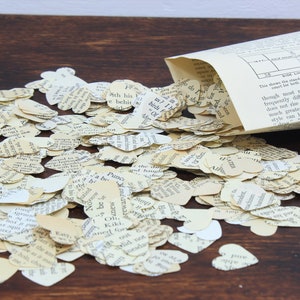 May include: A pile of heart-shaped confetti made from recycled book pages. The confetti is scattered on a wooden surface and spilling out of a paper cone. The text on the pages is visible, including the words "Ath his", "behir", "Panc", "rewar", "O", "Kiki", and "Of".