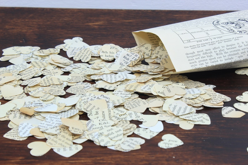 May include: A pile of heart-shaped confetti made from recycled book pages. The confetti is scattered on a wooden surface and spilling out of a paper cone. The text on the pages is visible, including the words "Ath his", "behir", "Panc", "rewar", "O", "Kiki", and "Of".