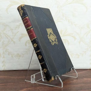 May include: Antique hardcover book titled "Captains Courageous" by Kipling. The book has a black cover with gold accents and a red spine. The book is displayed on a clear acrylic stand.