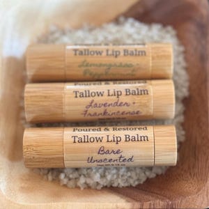 May include: Three wooden lip balm tubes with labels that read "Tallow Lip Balm". The top tube is labeled "Lemongrass Peppermint", the middle tube is labeled "Lavender Frankincense", and the bottom tube is labeled "Bare Unscented".