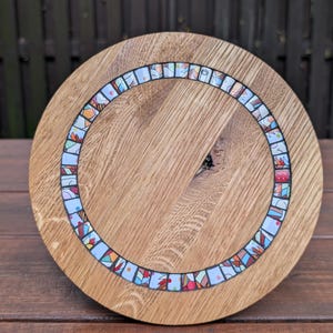 Può includere: Un tagliere rotondo in legno con un bordo a mosaico. Il tagliere è realizzato in legno marrone chiaro con un motivo a venature naturali. Il bordo a mosaico presenta disegni colorati e astratti.