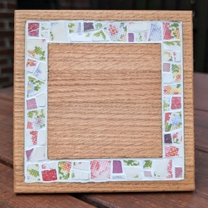 Può includere: Una cornice quadrata in legno con un bordo a mosaico. Il mosaico è composto da piccole tessere colorate con motivi floreali e geometrici. L'interno della cornice è in legno marrone chiaro.