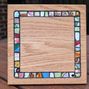 May include: A square wooden coaster with a mosaic tile border. The mosaic border features a variety of colourful tiles in shades of red, green, blue, and yellow. The coaster has a natural wood grain finish.