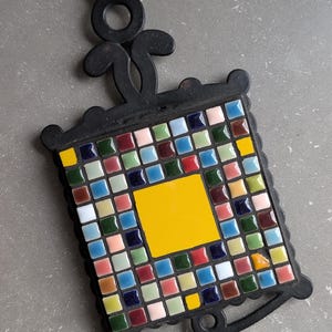 May include: Black metal trivet with a square mosaic tile design. The mosaic features a yellow square centre surrounded by small, colourful tiles in shades of red, blue, green, and pink. The trivet has a decorative handle.