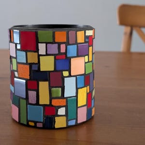 Puede incluir: Un jarrón cilíndrico de mosaico con interior y exterior negros. El mosaico está compuesto por varias baldosas cuadradas y rectangulares de colores en tonos rojos, azules, verdes, amarillos y morados. El jarrón está sobre una superficie de madera.