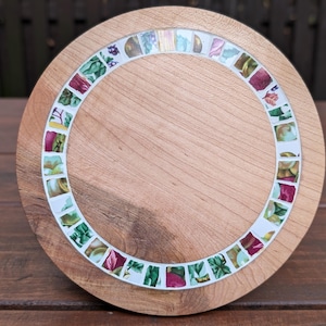 Può includere: Un vassoio rotondo in legno con un bordo a mosaico. Il mosaico presenta piccole tessere colorate con motivi floreali nei toni del verde, rosa e bianco. Il vassoio è in legno chiaro.