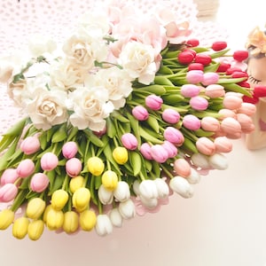 May include: A bouquet of colorful tulips and white roses with green stems. The tulips are in shades of pink, yellow, red, and white. A decorative figurine of a woman with a floral crown is on the right.