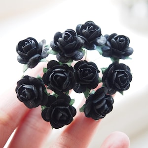 May include: A cluster of small, black paper roses with green stems. The roses are arranged in a circular pattern and are held in a hand.