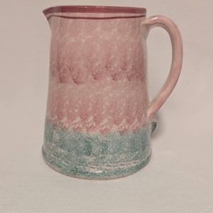 May include: A ceramic pitcher with a handle, featuring a speckled design. The top and handle are a solid pink, the middle section is pink with white speckles, and the bottom is teal with white speckles. The pitcher has a spout for pouring.