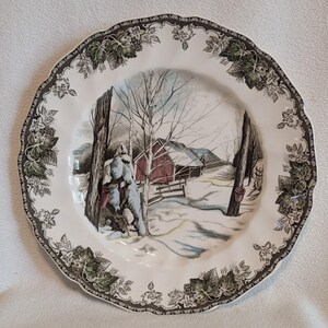 May include: A round, white ceramic plate with a winter scene featuring a red barn, snow, and leafless trees. The plate has a decorative border with green leaves and floral patterns.