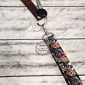 May include: A black and white key chain with a skull and floral design. The key chain is attached to a brown leather key fob with the text "Salem LV" on it. The key chain is attached to a silver key ring with two keys.