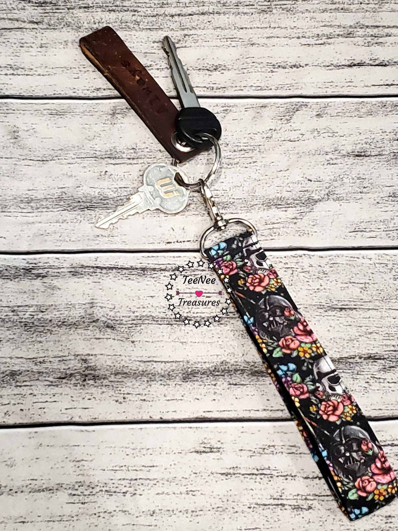 May include: A black and white key chain with a skull and floral design. The key chain is attached to a brown leather key fob with the text "Salem LV" on it. The key chain is attached to a silver key ring with two keys.