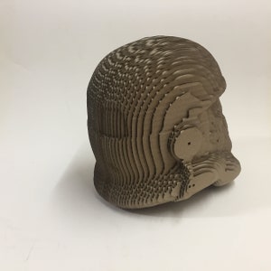 May include: A brown cardboard helmet with a layered design. The helmet has a textured surface and is made of multiple pieces of cardboard.
