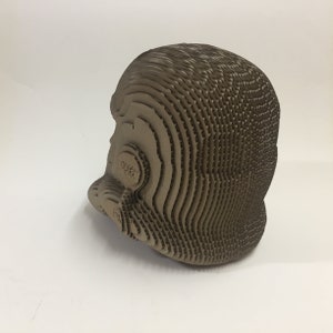 May include: A brown cardboard helmet with a layered design. The helmet has a textured surface and is made of multiple pieces of cardboard.