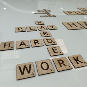 May include: Wooden letter tiles spelling out the phrase "Play Hard Work" on a white surface. The tiles are arranged in a grid pattern, with the letters "H", "A", "R", "D", "W", "O", "R", and "K" forming the words "Hard Work". The letters "P", "L", "A", "Y" are arranged above the word "Hard" to form the word "Play".
