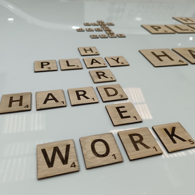 May include: Wooden letter tiles spelling out the phrase "Play Hard Work" on a white surface. The tiles are arranged in a grid pattern, with the letters "H", "A", "R", "D", "W", "O", "R", and "K" forming the words "Hard Work". The letters "P", "L", "A", "Y" are arranged above the word "Hard" to form the word "Play".