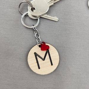 Può includere: Portachiavi in legno con la lettera M e un ciondolo a forma di cuore rosso. Il portachiavi è attaccato a un portachiavi d'argento.
