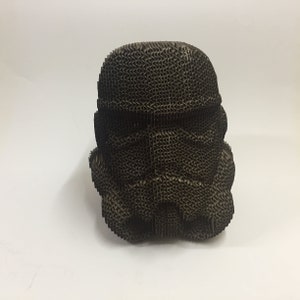 May include: A cardboard sculpture of a Star Wars Stormtrooper helmet. The helmet is made of brown corrugated cardboard and is detailed with the iconic features of the Stormtrooper helmet.