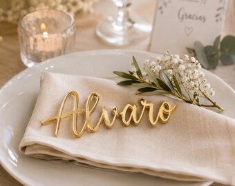 May include: A gold-colored name place card featuring the name Alvaro in a cursive script font, resting on a folded beige fabric napkin placed on a white ceramic plate. The name is rendered in a shiny, metallic finish.