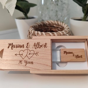 Peut inclure: Une boîte en bois contenant une clé USB. La boîte est gravée avec le texte "Marian & Albert 20 Ans" et deux cœurs avec des flèches qui les traversent.