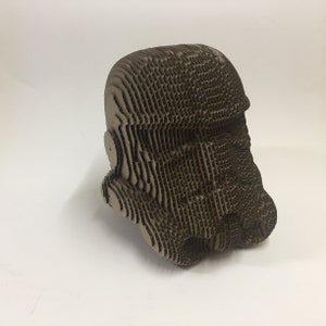 May include: A cardboard model of a Star Wars Stormtrooper helmet. The helmet is made of many layers of cardboard, creating a textured and detailed look.