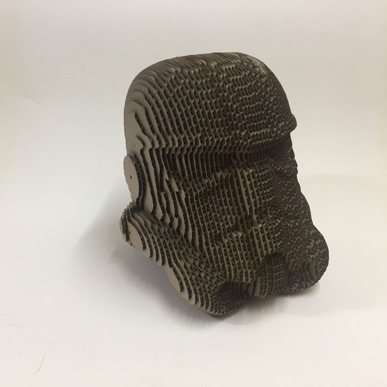 May include: A cardboard model of a Star Wars Stormtrooper helmet. The helmet is made of many layers of cardboard, creating a textured and detailed look.