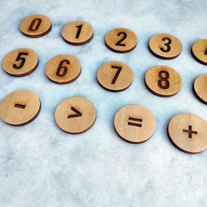 Può includere: Dischi di legno con numeri e simboli matematici. I dischi sono marrone chiaro con numeri e simboli marrone scuro. Sono presenti i numeri da 0 a 9, insieme ai simboli di addizione, sottrazione, uguale e maggiore di.