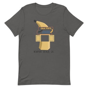 May include: A dark grey t-shirt featuring a graphic of a yellow banana and a sign that reads "Bluth's Original Frozen Banana" and "Newport Beach, CA."