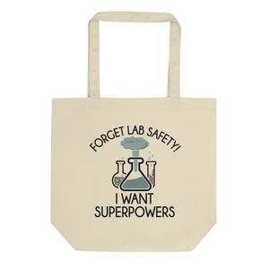 May include: A beige tote bag featuring a black graphic that reads "Forget Lab Safety! I Want Superpowers." The graphic includes a cartoon image of three beakers and a small explosion.