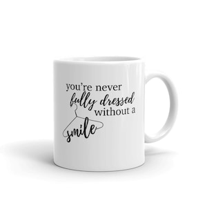 Puede incluir: Taza de cerámica blanca con un diseño de texto negro. El texto dice "you're never fully dressed without a smile" con un gráfico de percha. La taza tiene un asa curva y es adecuada para bebidas frías o calientes.