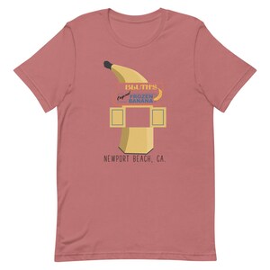 May include: A pink t-shirt featuring a graphic of a yellow banana and a sign that reads "Bluth's Original Frozen Banana, Newport Beach, CA."