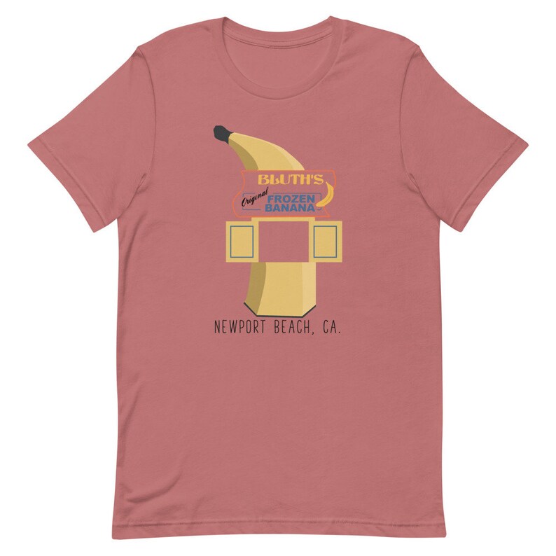 May include: A pink t-shirt featuring a graphic of a yellow banana and a sign that reads "Bluth's Original Frozen Banana, Newport Beach, CA."