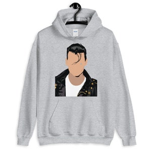 May include: A light gray hooded sweatshirt featuring a graphic of a person with black hair and a black leather jacket. The graphic has a minimalist design with a tear drop. The sweatshirt is hanging on a black hanger.