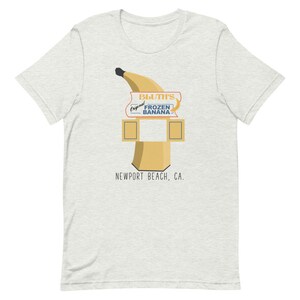 May include: A light grey t-shirt featuring a graphic of a yellow banana and a sign that reads "Bluth's Original Frozen Banana" and "Newport Beach, CA."