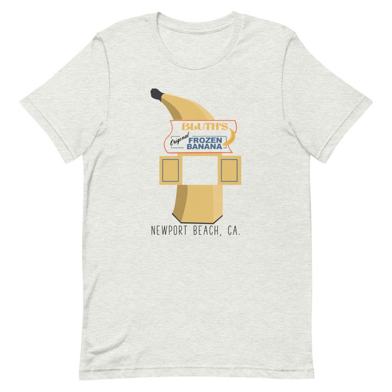May include: A light grey t-shirt featuring a graphic of a yellow banana and a sign that reads "Bluth's Original Frozen Banana" and "Newport Beach, CA."