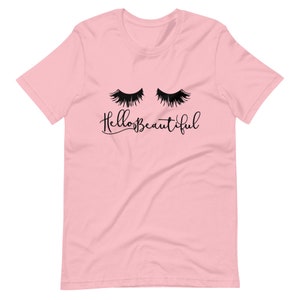 Puede incluir: Camiseta rosa claro con un gráfico negro de pestañas cerradas y el texto "Hello Beautiful". La camiseta tiene mangas cortas, cuello redondo y está hecha de una tela suave.