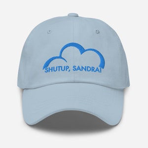 Può includere: Un berretto da baseball blu chiaro con una nuvola ricamata in blu e il testo "SHUTUP, SANDRA!"