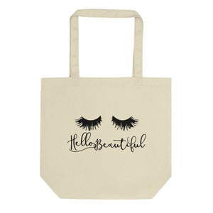 Puede incluir: Una bolsa de tela beige con asas negras y un gráfico negro de pestañas y el texto "Hello Beautiful".