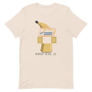 May include: A beige t-shirt featuring a graphic of a yellow banana and a sign that reads "Bluth's Original Frozen Banana" with the text "Newport Beach, CA."